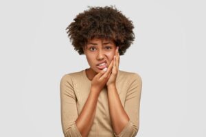 Woman with brown hair in a tan shirt holding two hands to her jaw in pain